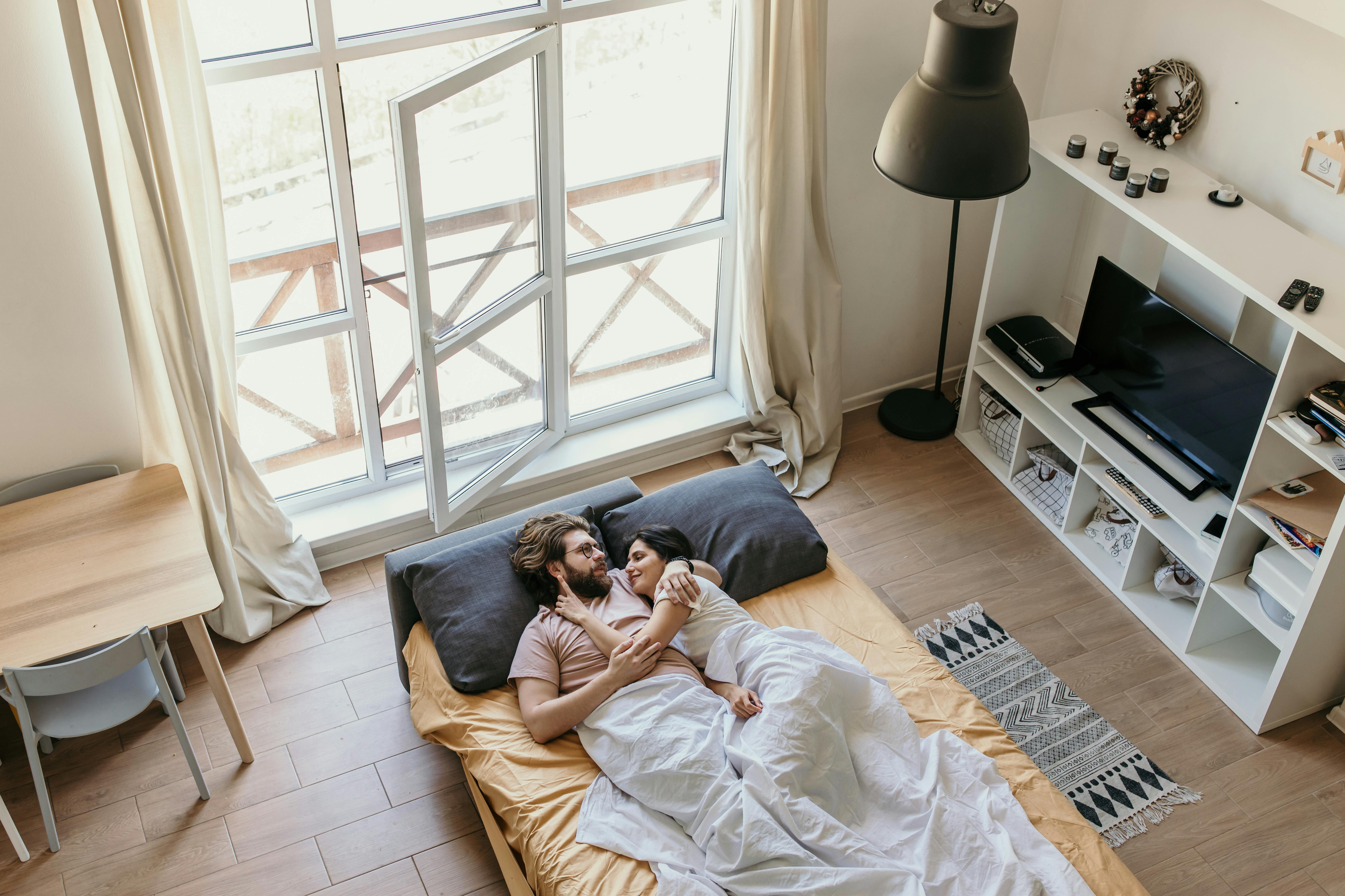A loving couple rests in bed by an open window, enjoying a peaceful morning together.