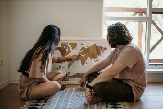 Couple sitting on floor pointing at a world map, planning their next travel adventure indoors.