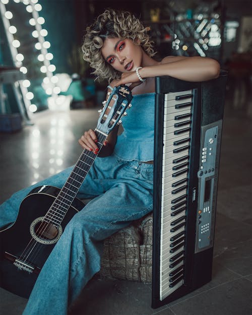 Free Confident young female musician with curly hair in trendy denim clothes sitting in modern studio with acoustic guitar and synthesizer during rehearsal Stock Photo
