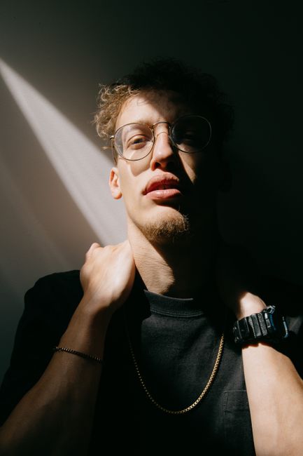 Serious young bearded guy in black t shirt and eyeglasses touching neck and looking at camera while standing against white wall