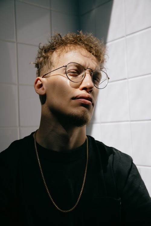 Focused young stylish unshaven male millennial in casual outfit and eyeglasses looking at camera thoughtfully against white tiled wall