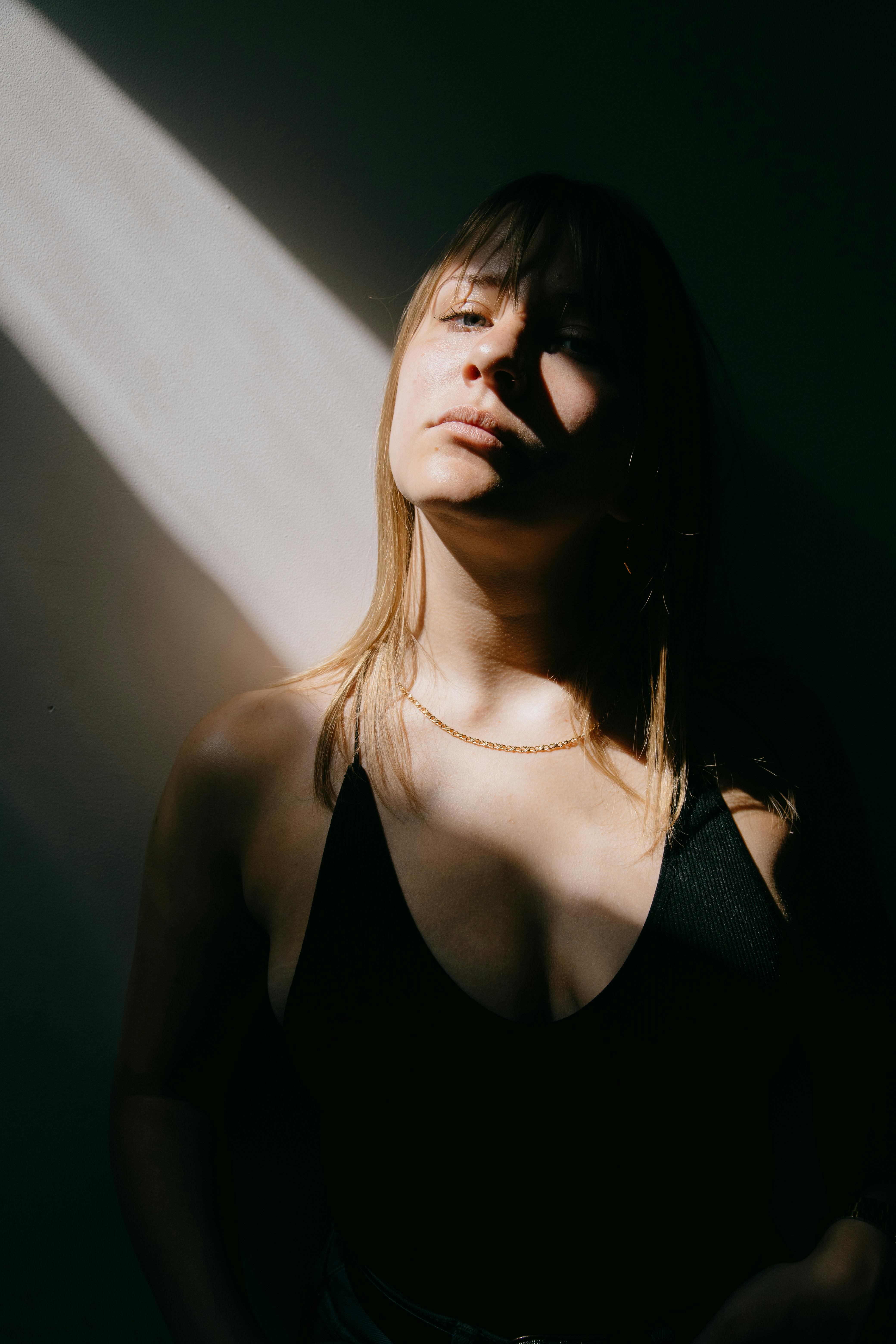 Sensual young female in black bodysuit standing near wall in dark room and looking at camera
