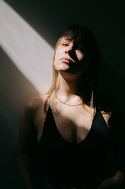 Sensual young female in black bodysuit standing near wall in dark room and looking at camera