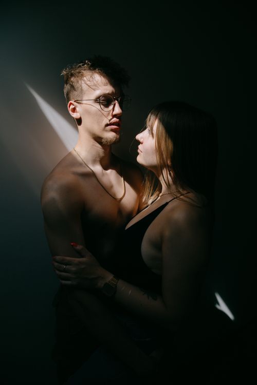 Free Side view of young shirtless man in eyeglasses hugging girlfriend near wall in dark room Stock Photo