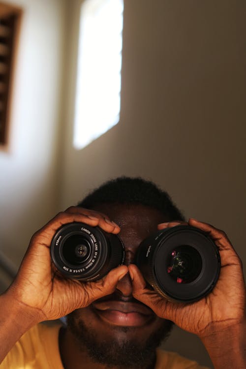 Unrecognizable African American male with short hair wearing yellow shirt looking through lenses of photo camera while spending time in room in daytime