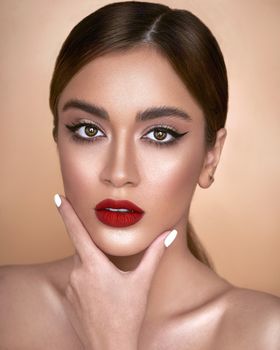 Portrait of young sensual female model with makeup touching face while looking at camera against light background