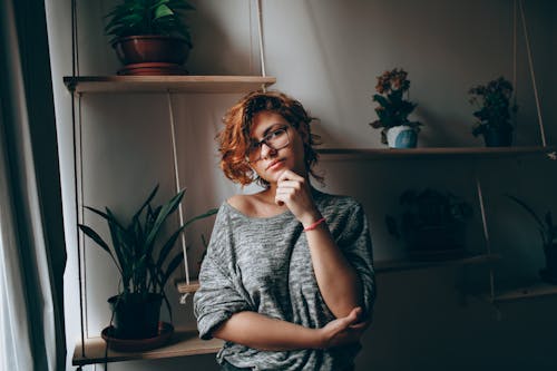 Young female in eyeglasses touching chin while thoughtfully looking at camera standing in room with plants on shelves