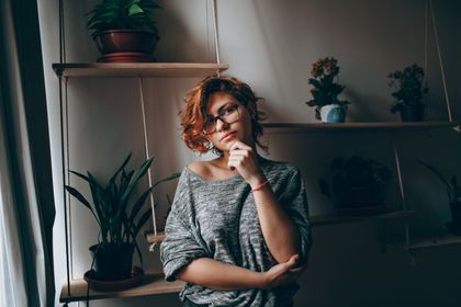 Young female in eyeglasses touching chin while thoughtfully looking at camera standing in room with plants on shelves — photo by Helena Lopes on Pexels