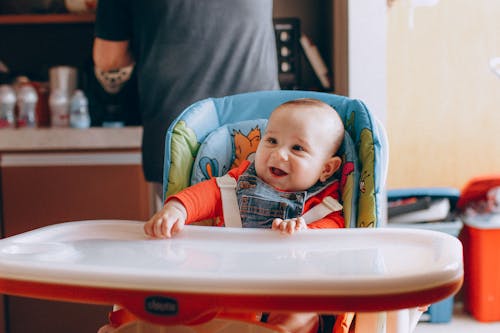 Adorable little baby in chair smiling brightly while waiting for food in kitchen