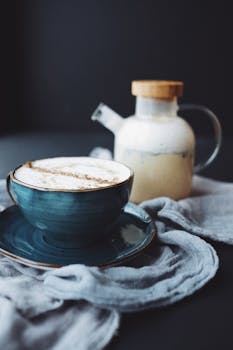 黑色背景前，一个蓝色陶瓷杯配着芳香的卡布奇诺，与玻璃水壶一同置于亚麻布上