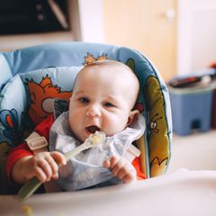 Baby with a spoon sitting strapped into a high chair with a padded seat printed with colourful cartoon animals