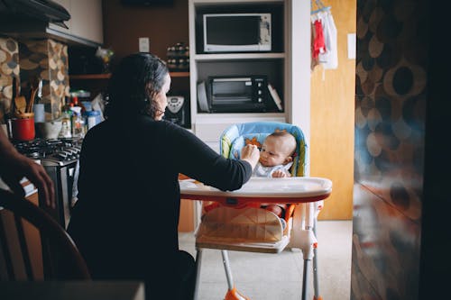 Small kid sitting in modern feeding chair eating food with nanny in cozy kitchen