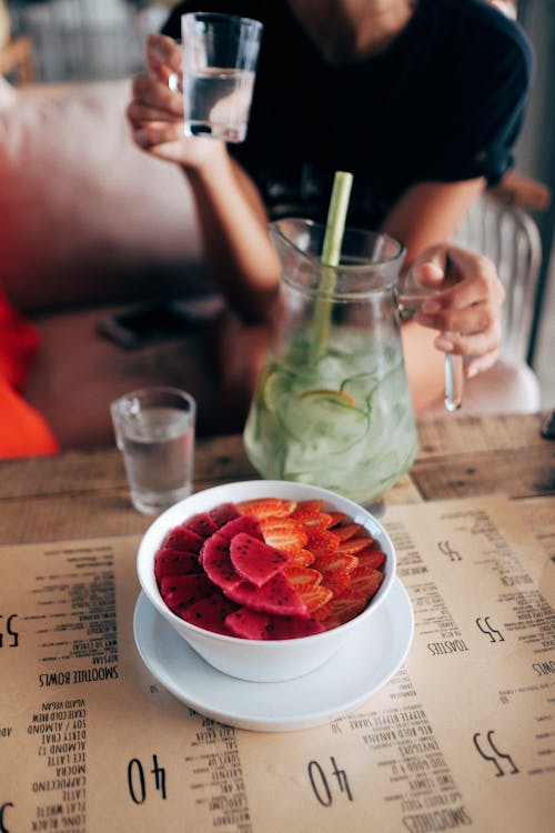 Free From above of healthy smoothie bowl decorated with fresh strawberries and dragon fruit and placed on table with anonymous woman drinking homemade lemonade on background Stock Photo