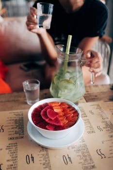 From above of healthy smoothie bowl decorated with fresh strawberries and dragon fruit and placed on table with anonymous woman drinking homemade lemonade on background