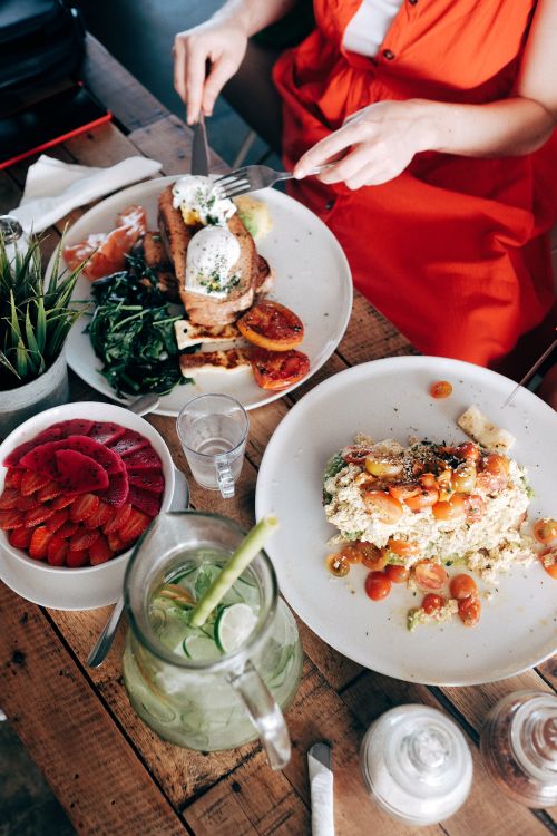 Free High angle of crop faceless young lady in casual dress eating toast with poached eggs sitting at table with salad smoothie bowl and lemonade Stock Photo