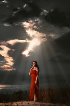 A woman in a red dress stands elegantly under a dramatic sunset in Rio's landscape.
