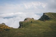 Person Standing on Green Grass Field Near Gray Rock Formation Under White Clouds
