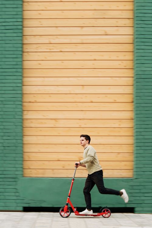 Free Man riding a red kick scooter against a modern urban backdrop, displaying a dynamic lifestyle. Stock Photo