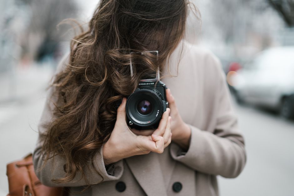 Unrecognizable young female photographer with long hair covering face using vintage camera while standing on street