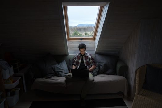 Concentrated young male freelancer in casual wear sitting on sofa in small attic room and working on laptop