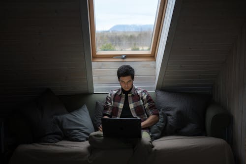 Serious young guy in checkered shirt sitting with laptop on couch while working remotely in cozy mansard room