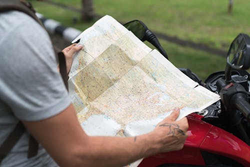Free Side view of crop unrecognizable male in casual clothes with backpack sitting on motorcycle while studying city map on street Stock Photo