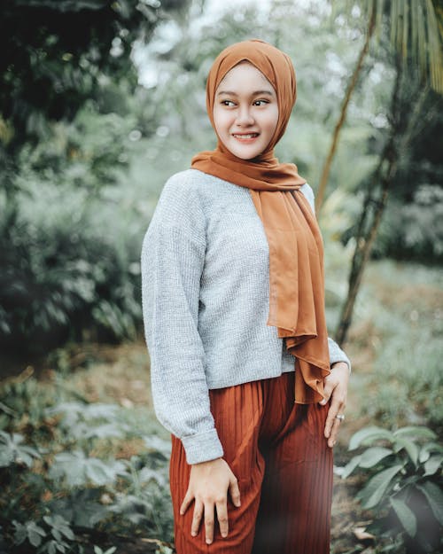 Happy young ethnic Muslim female in casual clothes and traditional headscarf standing in tropical garden and looking away