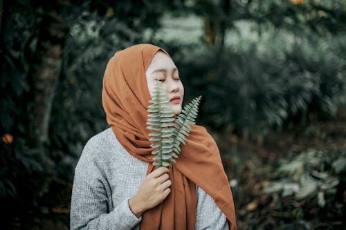 Pensive young Asian lady in hijab relaxing in tropical forest and smelling fresh leaves with closed eyes