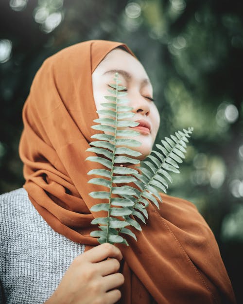 Low angle of peaceful young ethnic female in traditional headscarf holding fresh green leaves in hand and enjoying fragrance with closed eyes