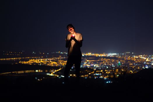 A man lights a cigarette, silhouetted against a cityscape at night, capturing a moody urban atmosphere.