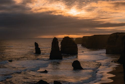 Majestic scenery of rocky formations in foamy ocean waving near sandy beach against picturesque sunset sky