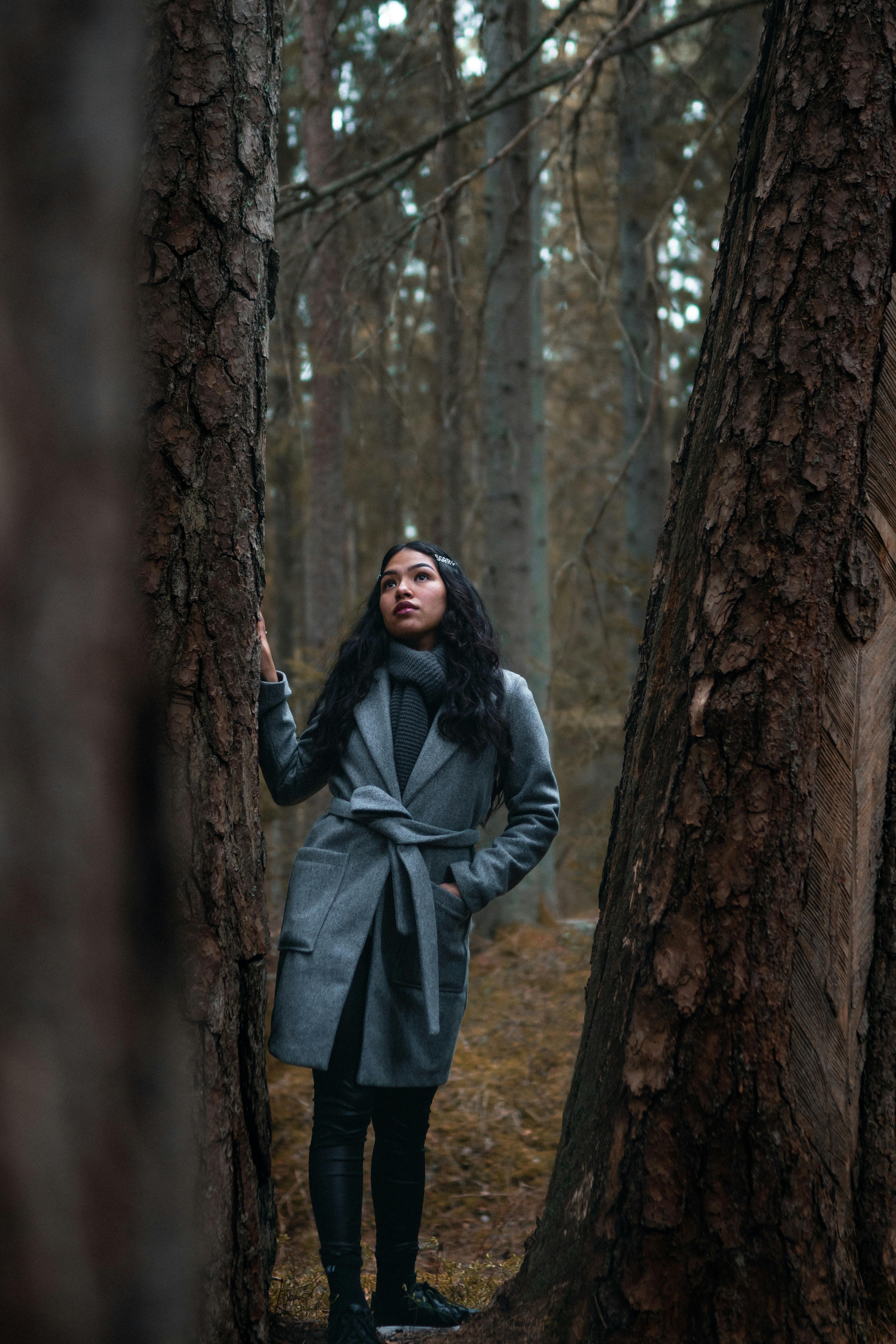 一名身穿灰色外套的女子，静静地站在森林的高大树木之间，显得孤独而自然