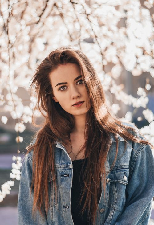 Free Pensive young beautiful female in black shirt and denim coat looking at camera while resting in shadow of blooming tree Stock Photo