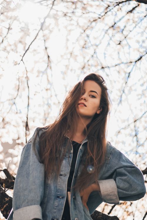 Free From below of serious young beautiful female with long hair in denim coat looking at camera while standing under tree with hand on waist in daylight Stock Photo