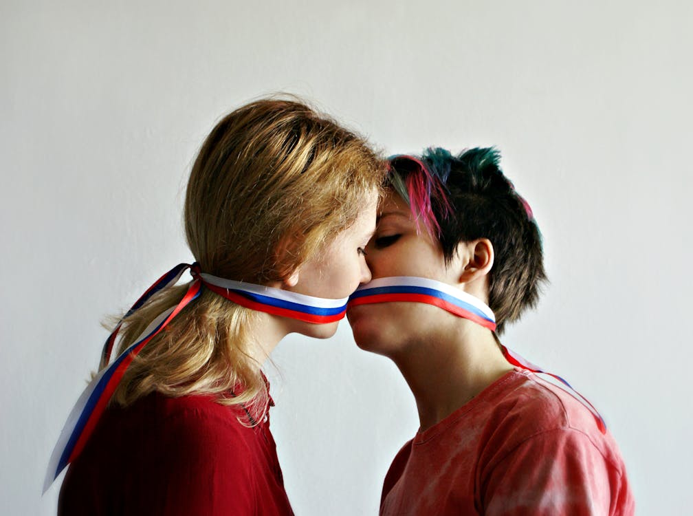 Free Side view of young female lovers in casual clothing looking at each other while kissing through ribbon in form of Russian flag on mouths Stock Photo