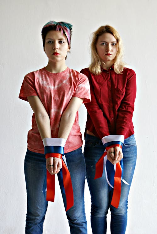 Free Pensive young slender females in casual clothing looking at camera while standing near grey wall with hands tied up with multi colored ribbons Stock Photo