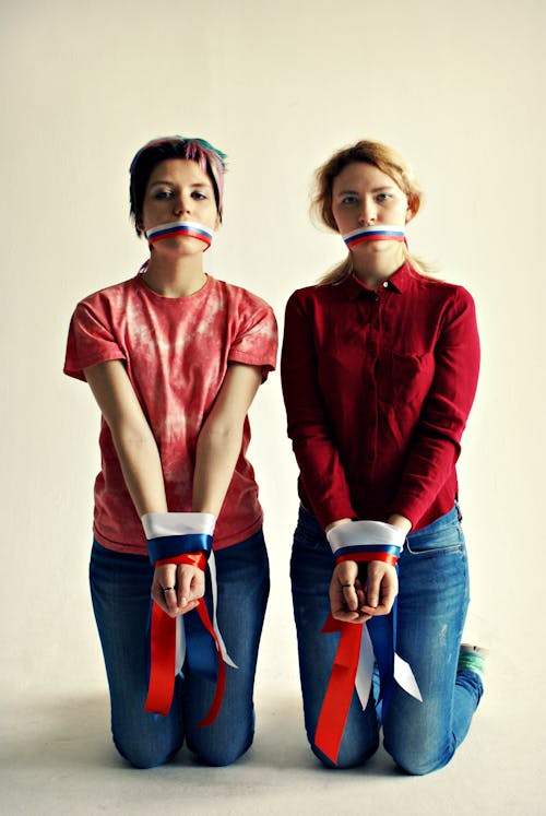 Full body of pensive young lesbian couple in casual clothing looking at camera while sitting on knees with ribbon on mouths and hands tied up next to grey wall