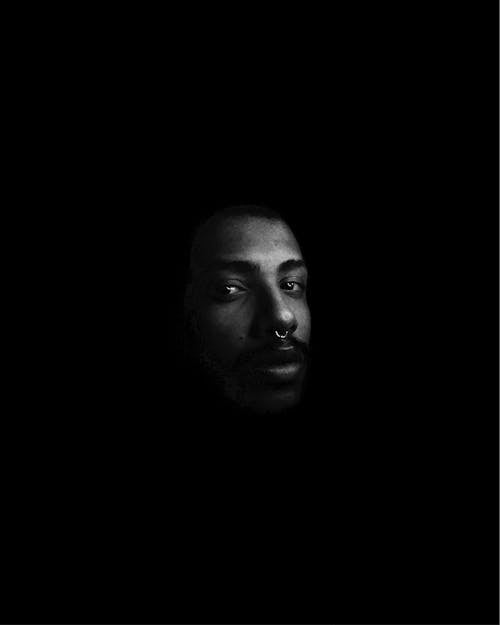 Black and white of pensive African American male with septum looking at camera in dark room