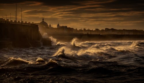 Dramatic scenery of stormy sea splashing near stony wall of waterfront of aged town at dusk