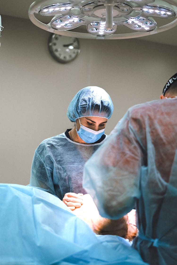 A Person Wearing Personnel Protective Equipment Holding A Patient Lying On Operating Table