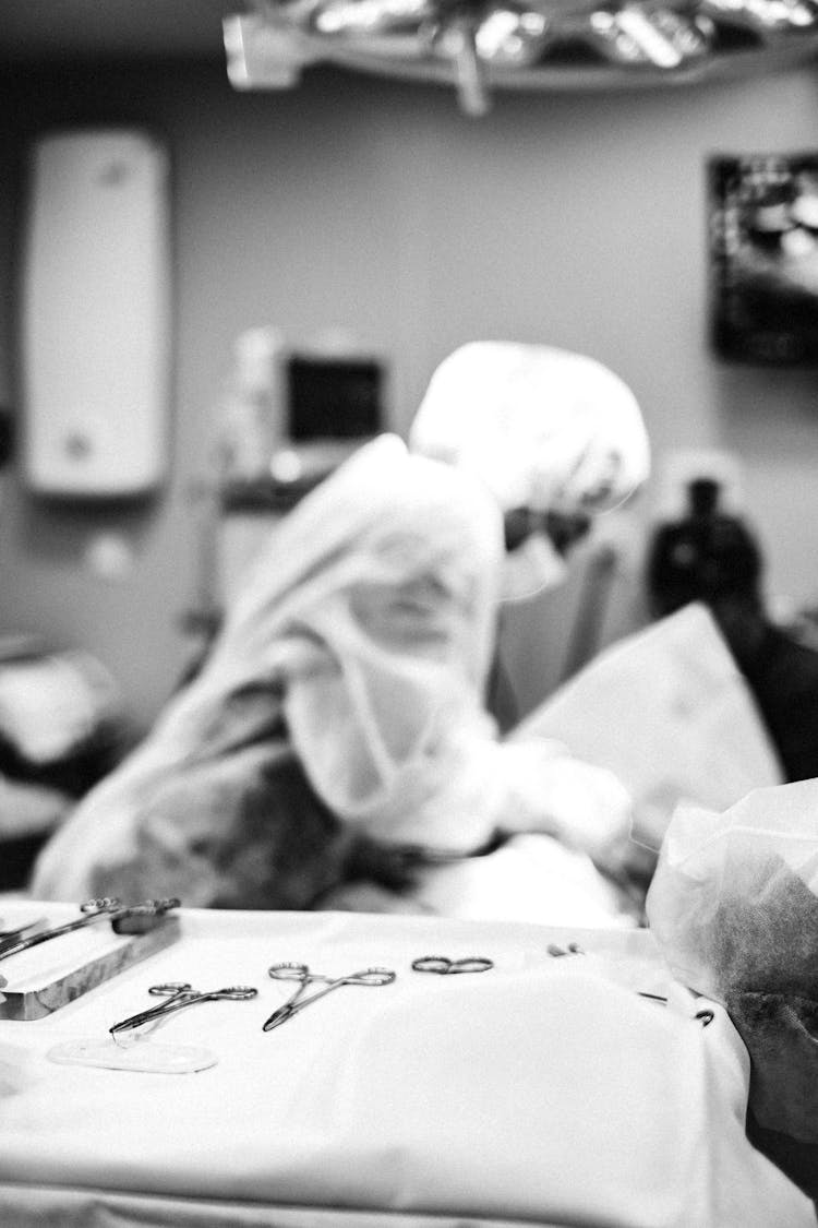 Grayscale Photo Of Surgical Instruments On Surface Near A Surgeon