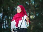 Low angle of young smiling woman in hijab holding mobile phone while standing against high trees