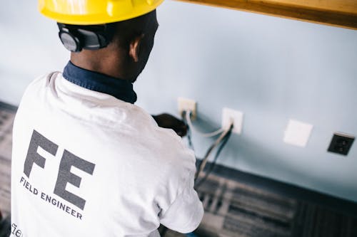 Free From above back view unrecognizable black field engineer wearing white shirt and protective hardhat sitting on floor and checking voltage in wall plugs Stock Photo