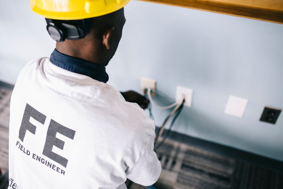 From above back view unrecognizable black field engineer wearing white shirt and protective hardhat sitting on floor and checking voltage in wall plugs .