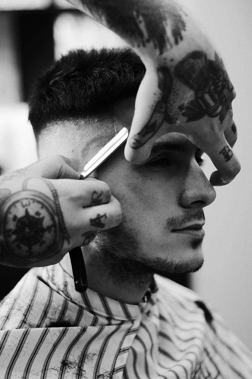 Free Black and white image showcasing a tattooed barber skillfully shaving a man's sideburns indoors. Stock Photo