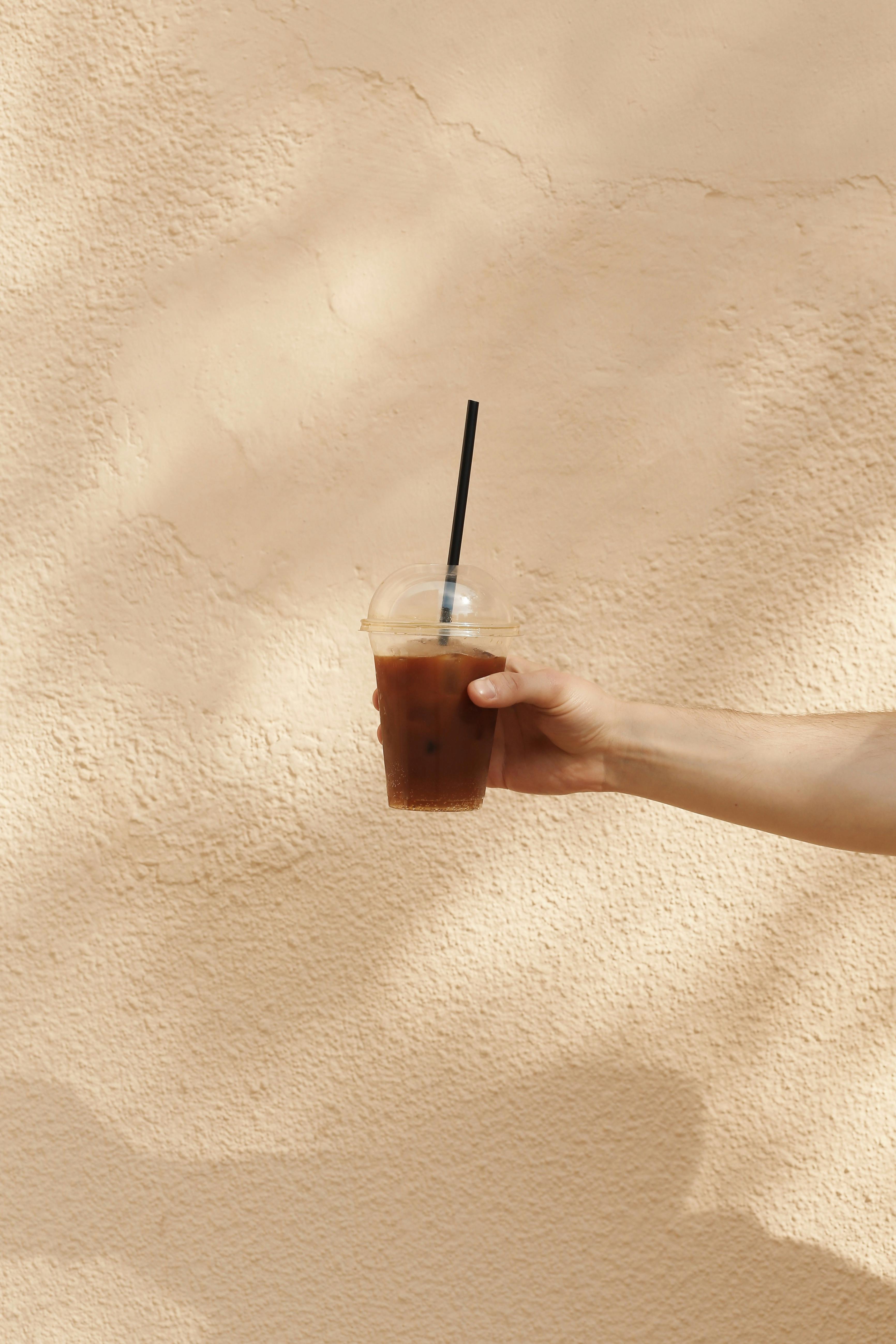 A person's hand holds an iced coffee in a plastic cup with a straw against a textured beige wall.