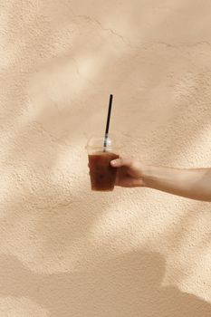 A person's hand holds an iced coffee in a plastic cup with a straw against a textured beige wall.