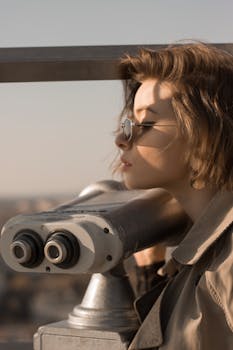 A fashionable woman in sunglasses uses binoculars to enjoy a city view.