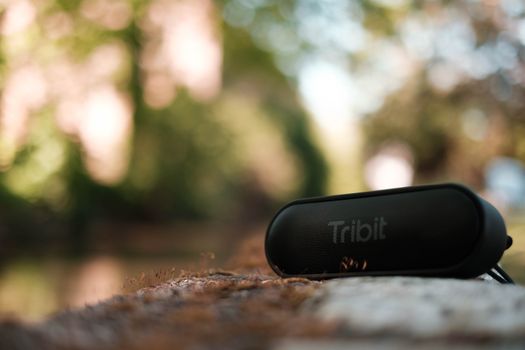 Contemporary black pocket speaker placed on mossy bench in lush park on sunny summer day