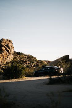 A black car parked on a desert road surrounded by rocky formations under a clear sky.